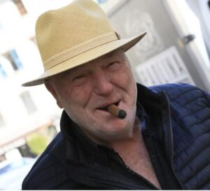 Manu Simonini, souriant, chapeau de paille et cigare aux lèvres, devant la vieille cave de Corte.